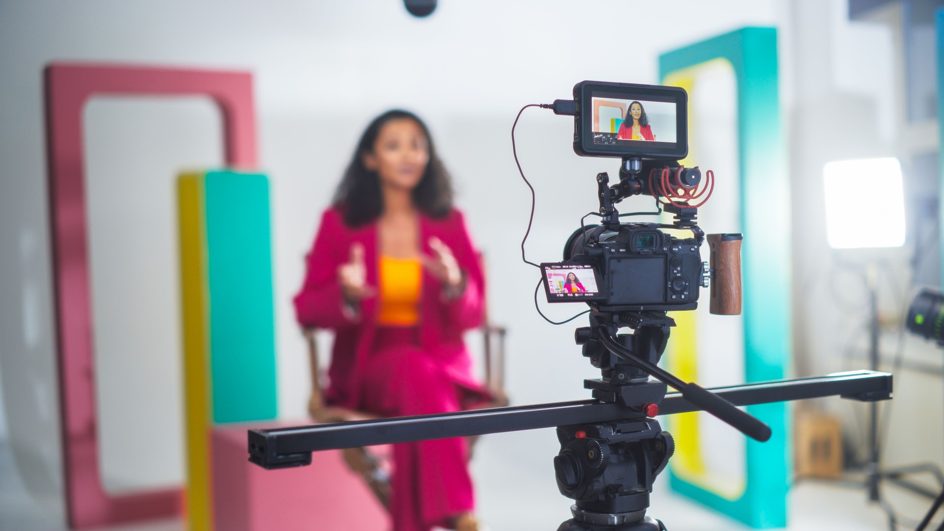 Vibrant Video Production Scene With A Black Female Host In A Pink Suit Engaging In A Lively Discussion, Captured On A Professional Camera Setup Against A Colorful, Modern Backdrop.